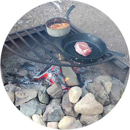 A campfire with a grill. Dinner is being prepared in a pot and skillet.